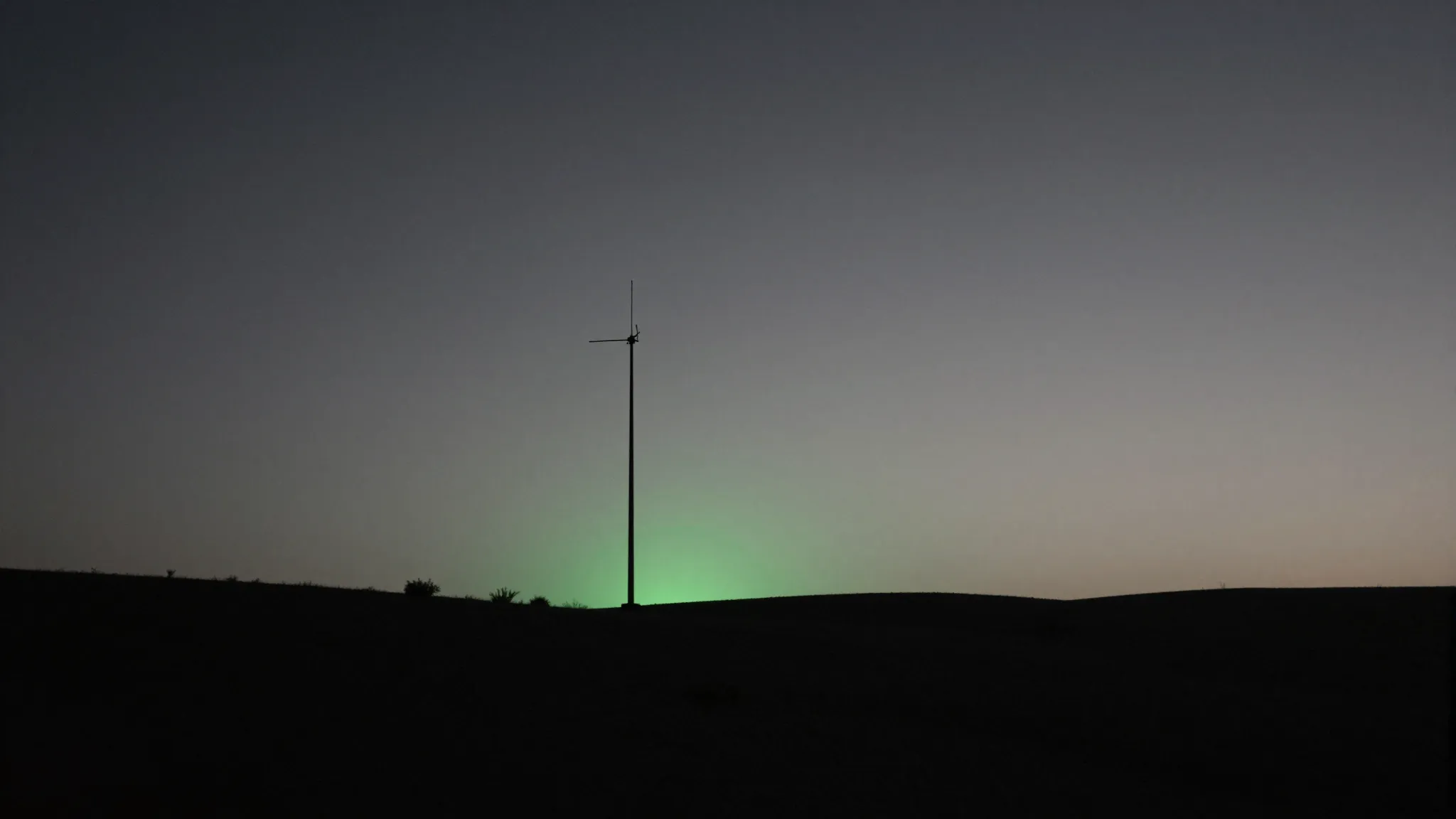 A lone antenna mast silhouetted on dark dusk terrain beneath a faint emerald horizon glow.